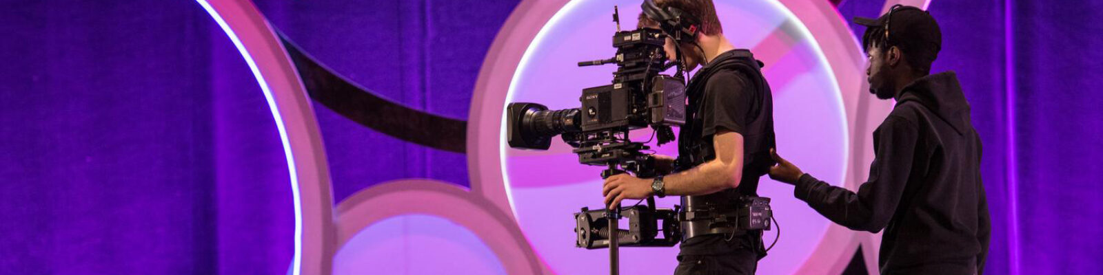 Two students handling a large recording camera on stage during the EVVY Awards, with a colorful purple backdrop behind them